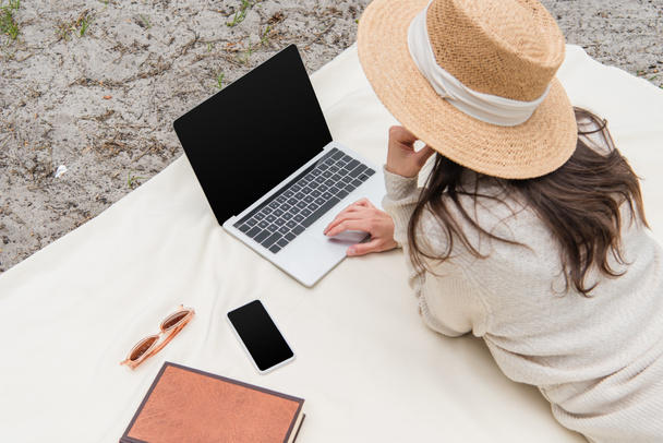 Hochwinkelaufnahme einer Frau mit Strohhut, die Laptop mit leerem Bildschirm benutzt, während sie auf einer Picknickdecke neben Smartphone, Sonnenbrille und Buch liegt  - Foto, Bild