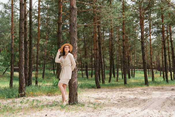 full length of cheerful young woman adjusting straw hat and standing in woods - Fotografie, Obrázek