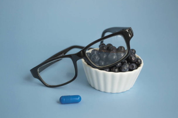 Conceito oftálmico de perto. Óculos, mirtilos e vitamina de mirtilo em um fundo azul. Produtos naturais para melhorar e manter a visão. Com espaço de cópia. - Foto, Imagem