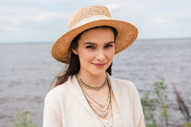 potěšená mladá žena ve slunečním klobouku při pohledu na kameru v blízkosti moře - Fotografie, Obrázek