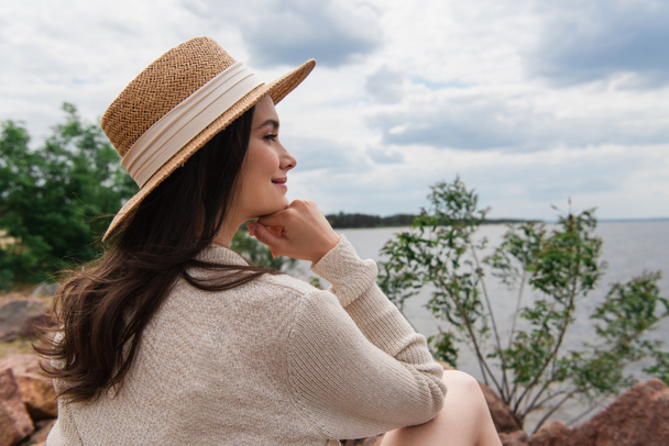 щаслива молода жінка в солом'яному капелюсі сидить біля моря
 - Фото, зображення