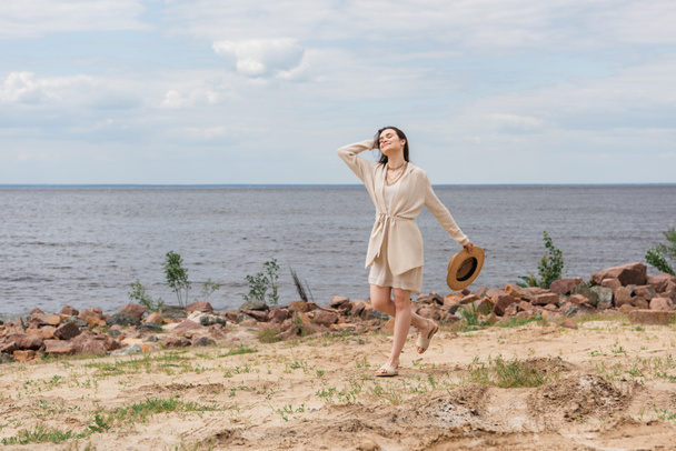 full length of happy young woman holding sun hat and walking near sea - Valokuva, kuva