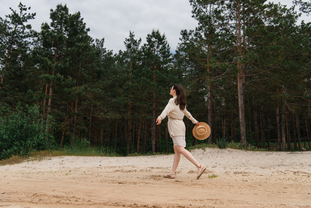 full length of brunette young woman in sunglasses holding straw hat and walking near forest  - Foto, Imagem
