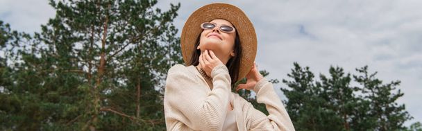 vue à angle bas de la jeune femme joyeuse dans les lunettes de soleil et chapeau de paille près de la forêt, bannière - Photo, image