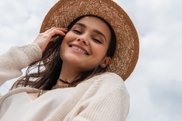 χαμηλή γωνία άποψη του χαρούμενη νεαρή γυναίκα σε ψάθινο καπέλο κοιτάζοντας την κάμερα - Φωτογραφία, εικόνα