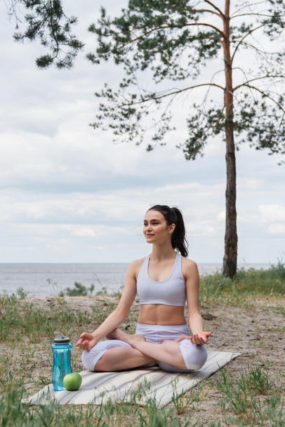 elégedett fiatal nő sportruházatban ül lótuszpózban és meditál jóga szőnyegen  - Fotó, kép