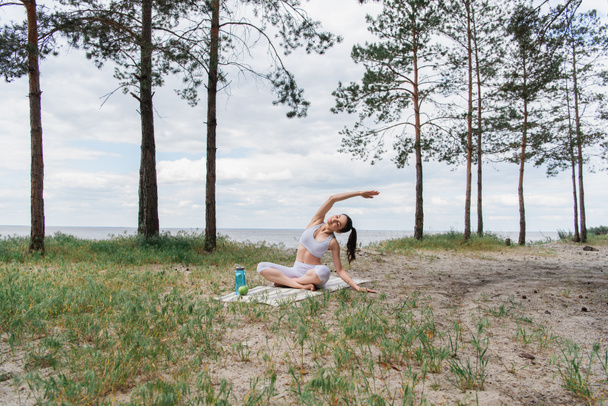morena mujer joven en ropa deportiva sentado en pose de loto y estiramiento en estera de yoga  - Foto, Imagen