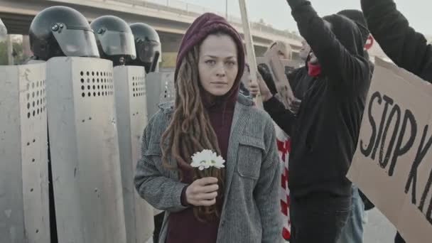 Portret van een jonge vrouw met dreadlocks die bloemen vasthoudt en naar de camera kijkt om te protesteren Mensen met borden die roepen voor onherkenbare oproerpolitie met schilden - Video