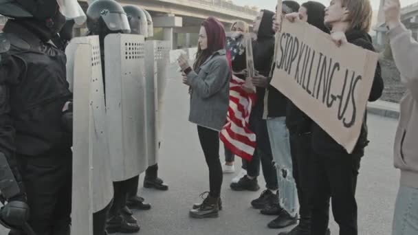 Slowmo średni strzał młodej kobiety z dredami wkładając kwiaty w tarcze policjantów zamieszek blokujących ulicę, podczas gdy młodzi ludzie ze znakami i flagą USA protestują i rozmawiają - Materiał filmowy, wideo