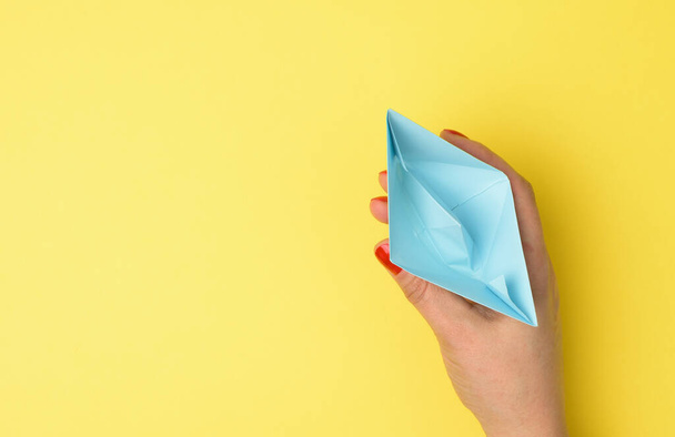 female hand hold a blue paper boat on a yellow background. Mentoring and support concept - Foto, afbeelding