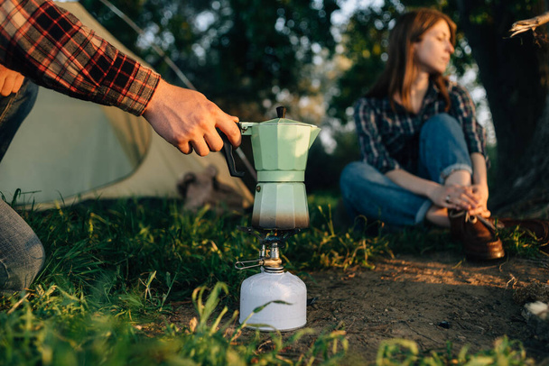 A human hand holds a geyser coffee maker the process of making coffee on a gas stove or burner in the forest. Camping cooking in nature. Lifestyle tourism and outdoor recreation. - Zdjęcie, obraz