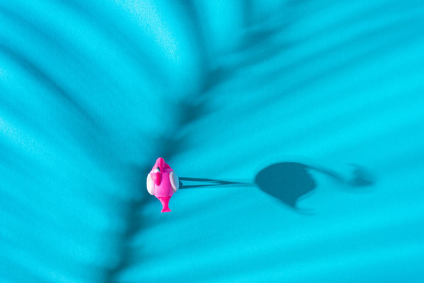 A flamingo toy standing on blue background with its sun shadow and palm leaf shadow. Drone shoot concept. Flat lay. - Photo, Image