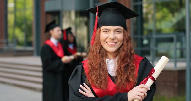 School graduate in academic dress and hat looking at the camera with happy smile. Adorable girl rejoicing near her university or school. Graduation concept - Zdjęcie, obraz