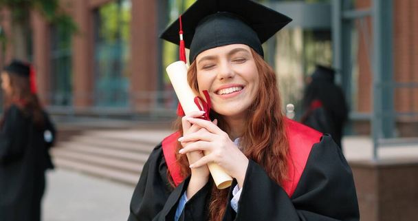 Educational concept. Happy caucasian student girl being happy at her diploma, graduation day. Ginger woman standing at the street with her friends at the background - Fotoğraf, Görsel
