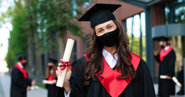 Portrait of caucasian female student wearing black hat and protective mask posing to the camera and showing her diploma after graduated from university during the covid 19 pandemic. Education concept - Foto, afbeelding