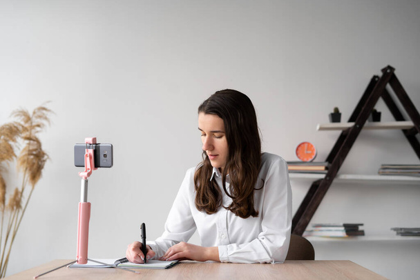 Συγκεντρωμένη φοιτήτρια κάθεται στο τραπέζι κρατά σημειώσεις σε ένα σημειωματάριο με ένα τηλέφωνο σε ένα τρίποδο. Το κορίτσι γράφει εργασίες από το κινητό της. Online εκπαίδευση και gadget. Θέση για κείμενο - Φωτογραφία, εικόνα