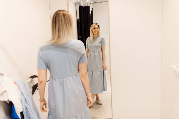 Charming blonde really likes herself in mirror offitting room in supermarket. Organization of sales process in supermarkets. Fashionable clothes. Shopping and pleasure. Convenience and comfort. Lifestyle. - Foto, immagini