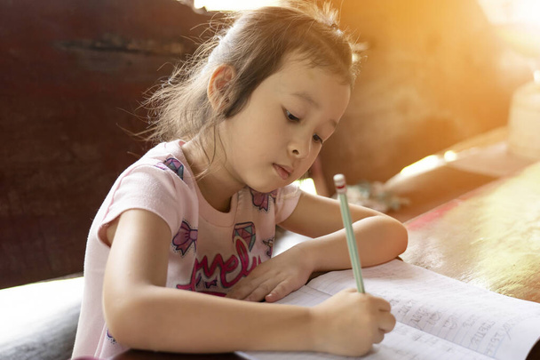 Asiatisches Mädchen sitzt zu Hause auf einem Schreibtisch und schreibt ein Buch. Online-Bildung. Neue normale Bildung. Lese- und Schreibkonzepte für Kinder. Bildung von zu Hause aus - Foto, Bild