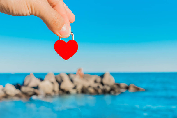 The man's hand holds a small red heart in the form of a castle against the backdrop of the sea coast and the stones of the breakwater on the horizon line. Symbol or sign of love. - Φωτογραφία, εικόνα