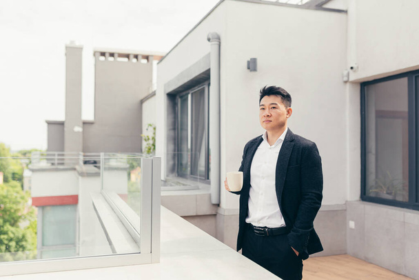 Young successful Asian businessman in a business suit drinking tea or coffee standing on the balcony or terrace of a modern office center - Photo, image