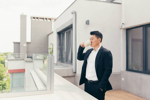 Young successful Asian businessman in a business suit drinking tea or coffee standing on the balcony or terrace of a modern office center - Foto, imagen