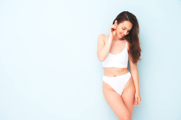 Portrait of young beautiful smiling woman in white lingerie. Sexy carefree cheerful model in underwear posing near light blue wall in studio. Positive and happy female enjoying morning - Φωτογραφία, εικόνα