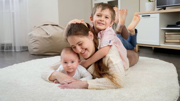 Rapaz bonito abraçando sua mãe e irmãozinho deitado no tapete na sala de estar. Paternidade, felicidade das crianças e relacionamento familiar - Foto, Imagem