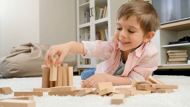 Kleiner Junge baut Haus aus Spielzeug-Holzklötzen auf dem Boden im Wohnzimmer. Konzept der Kindererziehung und Spiele zu Hause - Foto, Bild