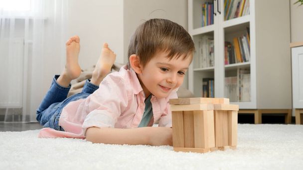 Lächelnder Junge, der auf dem Boden liegt und Haus oder Turm aus Holzklötzen baut. Konzept der Kindererziehung und Spiele zu Hause - Foto, Bild