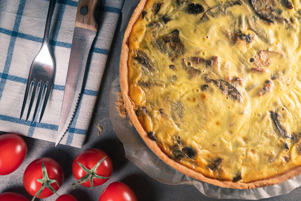 Top view of a freshly baked yellow quiche with mushrooms still on a baking paper, sitting on a dark kitchen table surface with cutlery set, tomatoes, mint leaves - Photo, Image