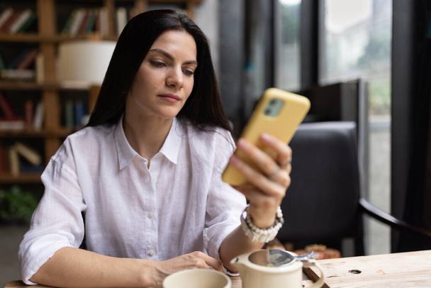 Glückliche junge kaukasische Millennial- oder Gen-Z-Frau mit langen brünetten Haaren, die mit dem Smartphone streamt und Social-Media-Blogs in modernen Cafés aufnimmt. Influencer nutzen soziale Netzwerke. - Foto, Bild