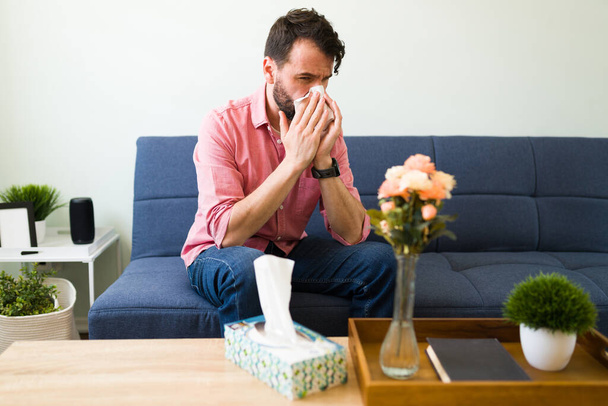 Adult man sick with a contagious virus sneezing while sitting in the living room. Latin man suffering from a bad cold blowing his nose - Photo, Image