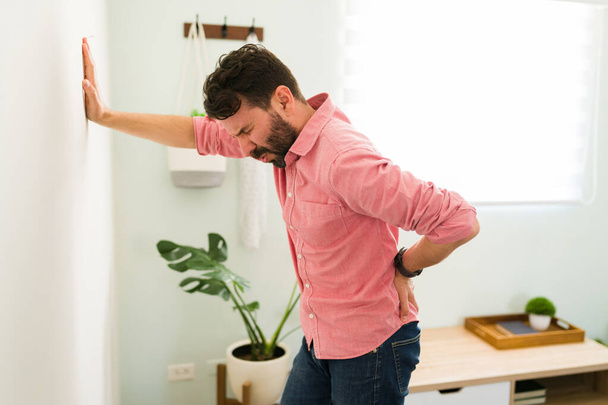 腰をこすり腰痛に苦しみながら、リビングの壁に寄りかかって息をのむストレスの多い男。 - 写真・画像