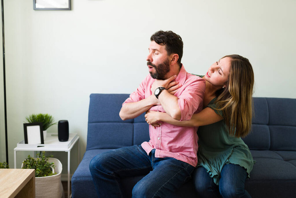 Stressed young woman doing the Heimlich maneuver on an hispanic man choking while sitting together on the couch  - Фото, изображение