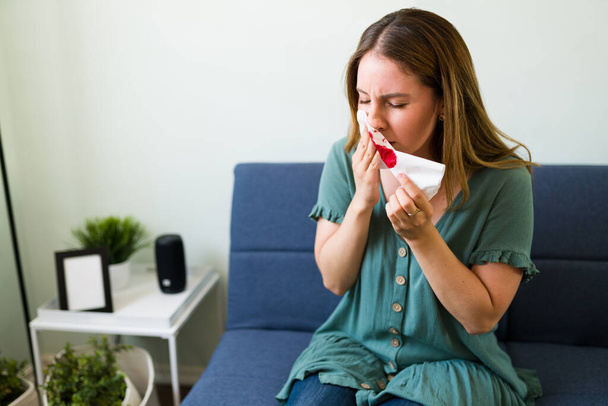 Młoda kobieta dmucha nos bibułą i krwawi z nosa siedząc samotnie w salonie - Zdjęcie, obraz