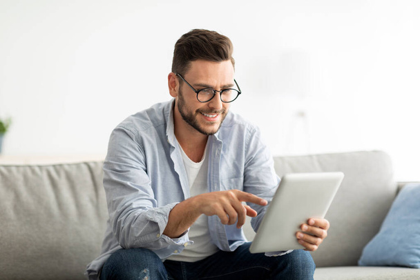 Freizeitkonzept. Glücklicher Mann mit digitalem Tablet, auf Couch im Wohnzimmer sitzen, Raum kopieren - Foto, Bild
