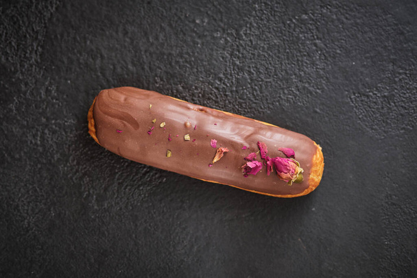 Eclair csokoládé cukormázzal, díszített szárított rózsa. Hagyományos francia desszert. Édességek az ünnepre egy sötét asztalon. - Fotó, kép
