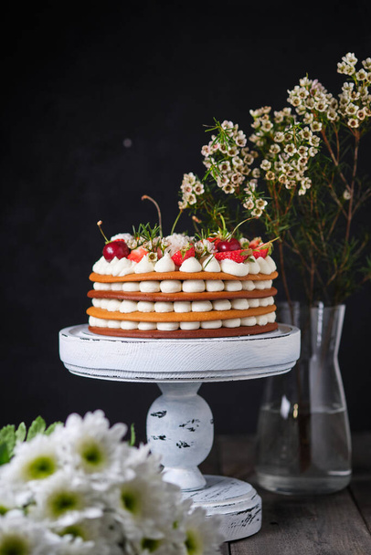 Honey cake with fresh berries, flowers and sweets. The layers of the cake are soaked in cream cheese cream. Holiday cake on a wooden stand. - Zdjęcie, obraz