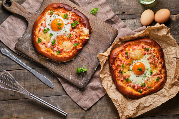 Rustikale Pizza auf Hefeteig mit Schinken, Käse, Ei, Tomatensauce und Kräutern. Appetitlich-schmackhaftes Essen auf einem Holztisch. - Foto, Bild