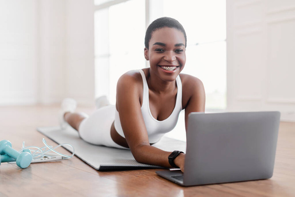 Young black woman lying on yoga mat using laptop - 写真・画像