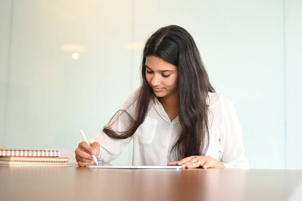 Tiro de una joven estudiante sentada en la mesa y usando lápiz lápiz lápiz de escritura en la tableta en el hogar moderno. - Foto, Imagen