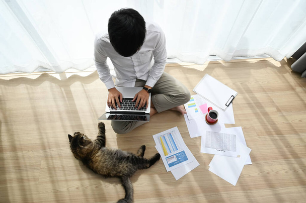 Młody mężczyzna siedzi na podłodze z kotem i pracuje na laptopie w domu. - Zdjęcie, obraz