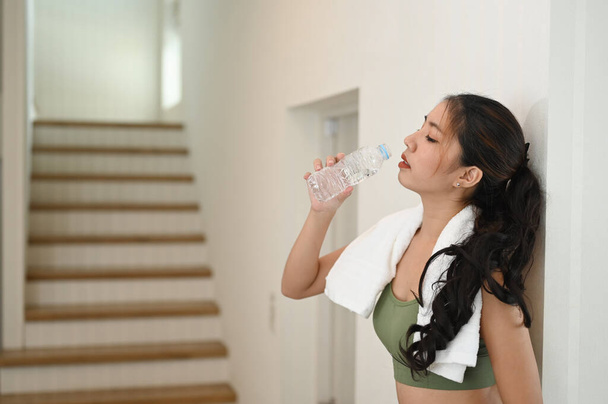 Красива фітнес жінка п'є воду з пляшки, стоячи у вітальні
. - Фото, зображення