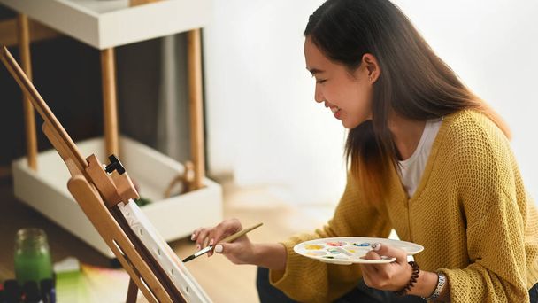 Tiro de artista feminina segurando pincel e colocando traços de tinta com cuidado ao pintar imagem no cavalete em seu estúdio. - Foto, Imagem