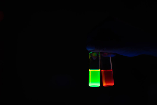 Una investigadora que sostiene viales de vidrio resplandeciente de reacción fotoquímica química en un laboratorio orgánico - radiactivo - fluorescencia. Un espacio de copia fondo negro. Laboratorio de química médica. - Foto, Imagen