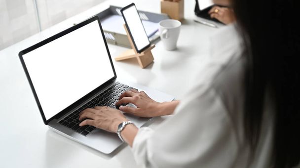 Przycięte zdjęcie bizneswoman pracującej na laptopie i smartfonie z białym ekranem w nowoczesnym biurze. - Zdjęcie, obraz