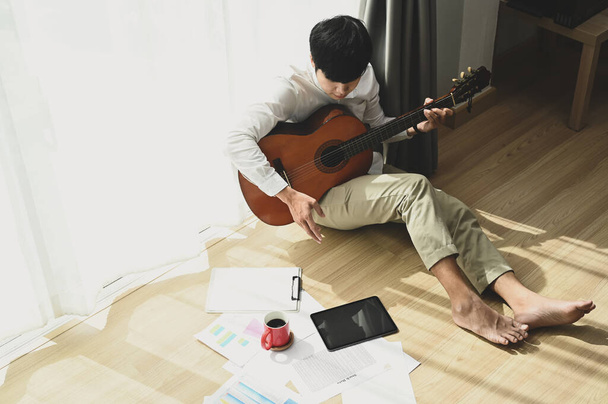 Junger Mann sitzt zu Hause am Fenster und spielt Akustikgitarre. - Foto, Bild