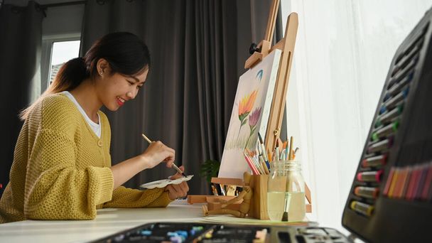 Portrait of happy young woman artist holding palette and paintbrush while working on painting in her workshop. - Photo, Image