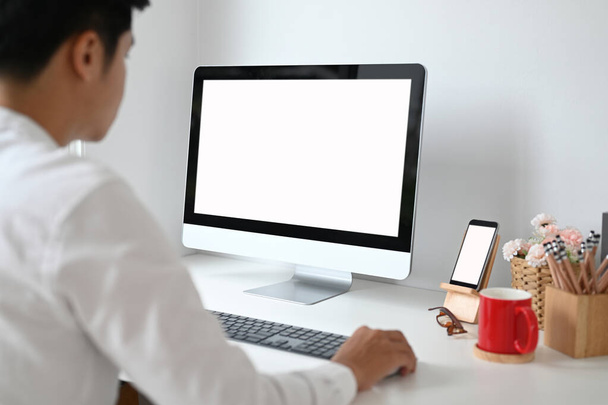 Στιγμιότυπο του νεαρού άνδρα να σχεδιάζει το έργο του στον υπολογιστή με λευκή οθόνη στο λευκό γραφείο. - Φωτογραφία, εικόνα