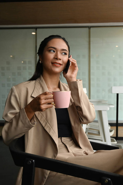 Shot of business young woman talking on mobile phone while holding coffee cup in the office. - Foto, Bild